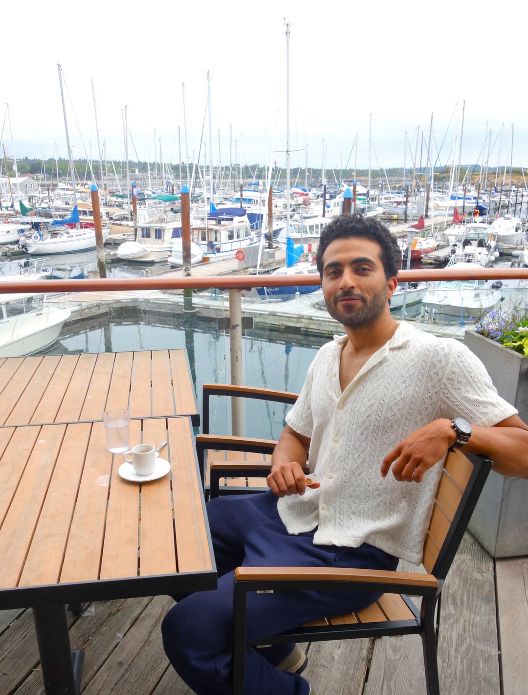 A man in a cream shirt sits at a marina patio table, sailboats moored behind him.
